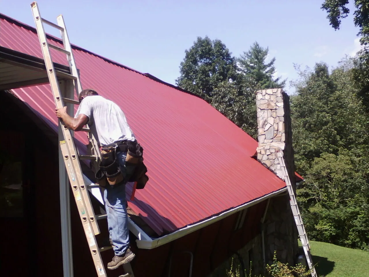 Roofer on a ladder beside a red metal roof and stone chimney — Soffit & Fascia Repair by Roofing Brothers in Decker